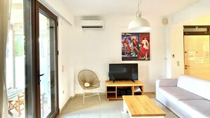 Uma sala de estar moderna com um sofá, uma mesa de centro de madeira, uma televisão e uma cadeira. Há uma porta de vidro deslizante que dá para uma área externa.