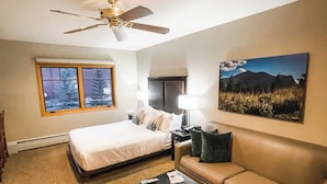 A hotel room with a bed, a sofa, a coffee table, a ceiling fan, a window with a view of trees, and a painting on the wall. - Grand Timber Lodge Studio Ski in/out (Breckenridge)