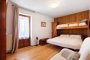 Una habitación con literas, suelo de madera, un radiador, una ventana con cortinas y un pequeño cofre de madera.