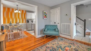 Una cocina moderna con armarios blancos, un comedor con mesa y sillas de madera, un sillón color turquesa y un tapete estampado.