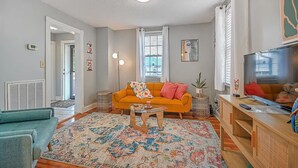 Una sala de estar con un sofá, una mesa de centro y un televisor. Hay una alfombra estampada en el suelo, y una ventana con persianas.