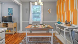 Un comedor con una mesa de madera dispuesta para cuatro, un televisor sobre un soporte y una ventana con persianas.