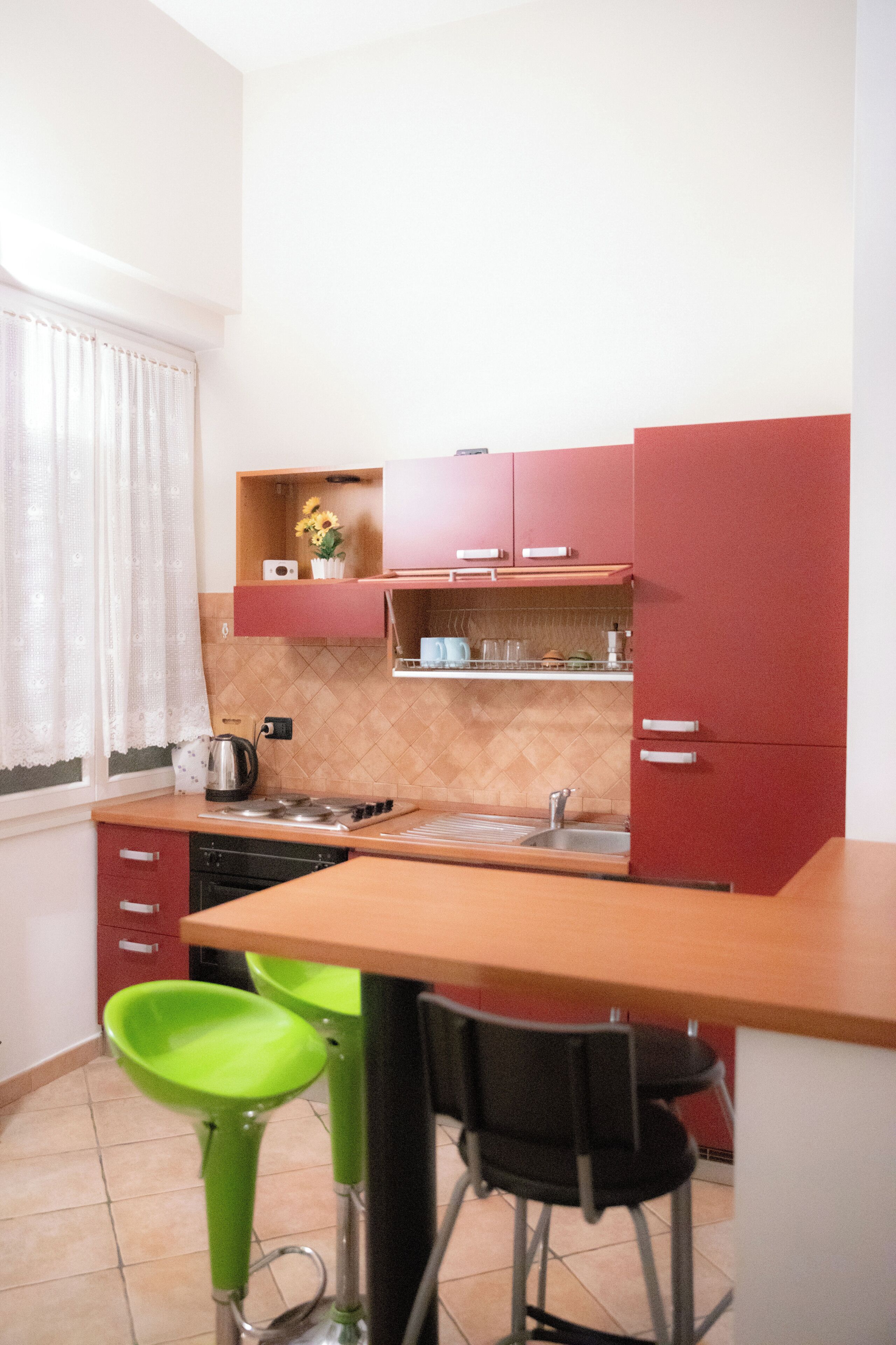 Cocina compacta con lavamanos, estufa y una pequeña encimera. Hay dos taburetes de bar y una ventana con cortinas.