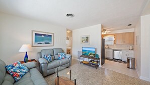 Sala de estar con sofá, televisor y una mesa de centro. Zona de cocina con armarios y un lavamanos.