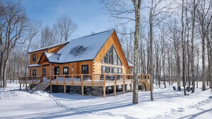 Une cabane en rondins avec une terrasse en bois et des fondations en pierre, entourée de neige et d’arbres.