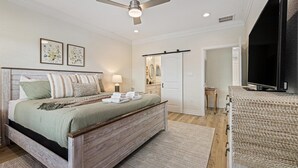A bedroom with a wooden bed, a ceiling fan, a television, and a door leading to a bathroom. - After Dune Delight Pointe San Luis far West End (Galveston)