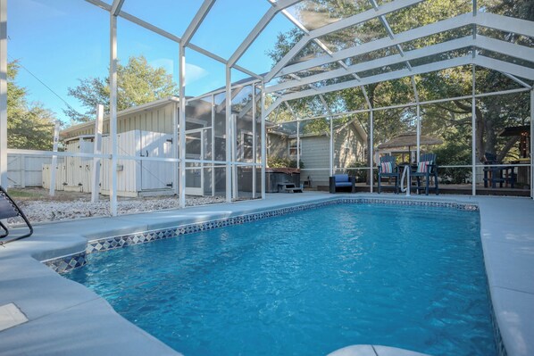 A pool area with a clear pool, a covered patio with chairs, and a white fence surrounding the property. - 2 Step: Pool, Hot Tub and Dog-Friendly (Oak Island)