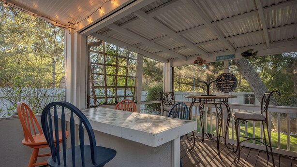 Una veranda coperta all'aperto con tavolo e sedie, un cartello con la scritta "Welcome to Oakley Backyard Paradise" e vista sugli alberi e il giardino circostanti.