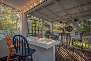 A covered outdoor patio with a table and chairs, a sign reading "Welcome to Oakley Backyard Paradise," and a view of the surrounding trees and yard. - 2 Step: Pool, Hot Tub and Dog-Friendly (Oak Island)