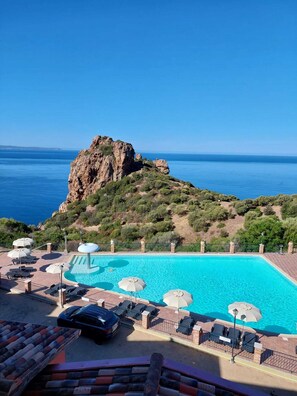 A pool area with lounge chairs and umbrellas, a car, and a view of the sea and a rocky hill. - Il Sole & La Luna (Nebida)