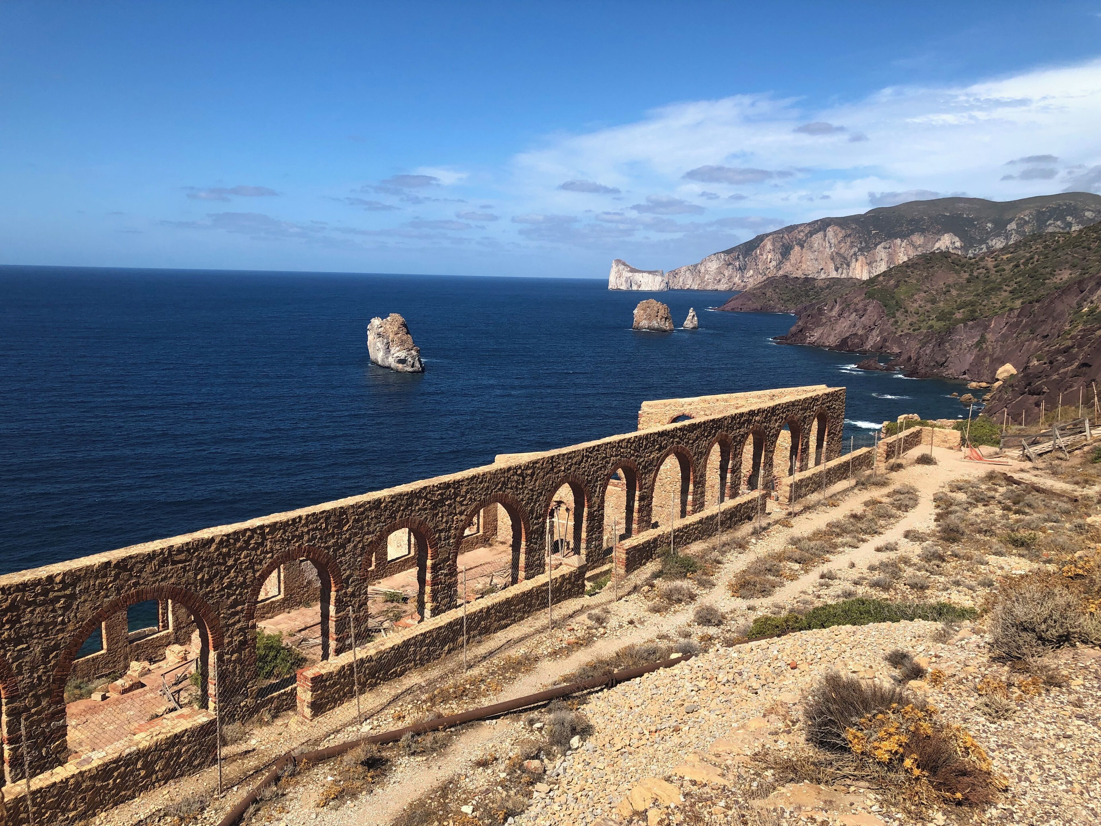 Un paisaje costero con un acueducto de piedra, afloramientos rocosos y una vista despejada del mar.