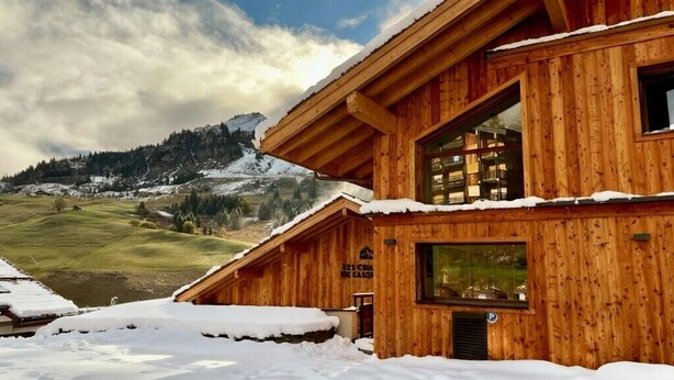 Un chalet en bois doté d’un balcon, de grandes fenêtres et offrant une vue sur un paysage montagneux enneigé.