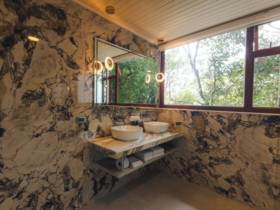 Un baño con paredes de mármol, un espejo grande y una ventana con vistas a árboles.