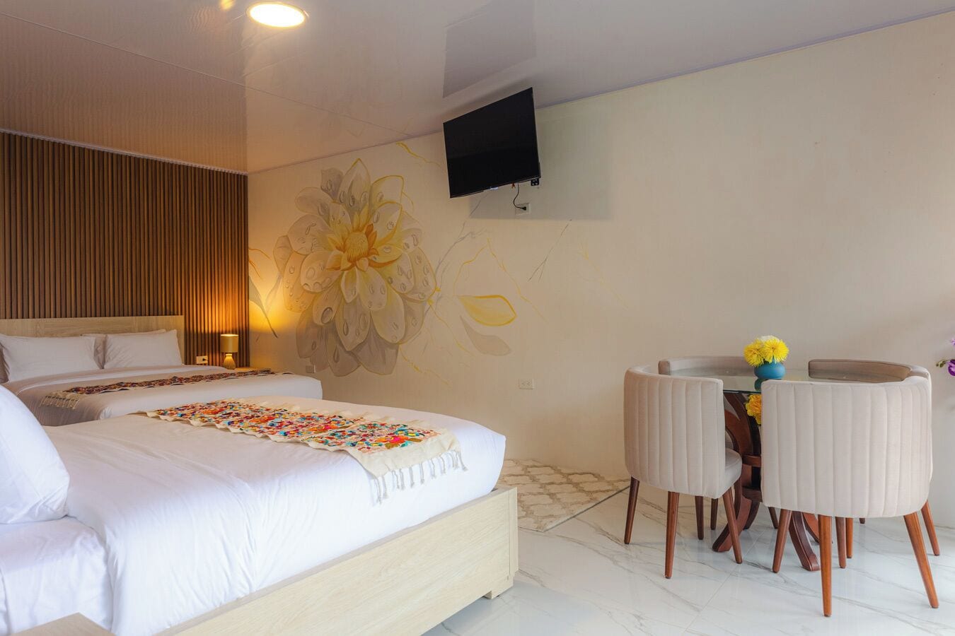 A hotel room with two beds, a wall-mounted TV, a desk with chairs, and a large floral mural.