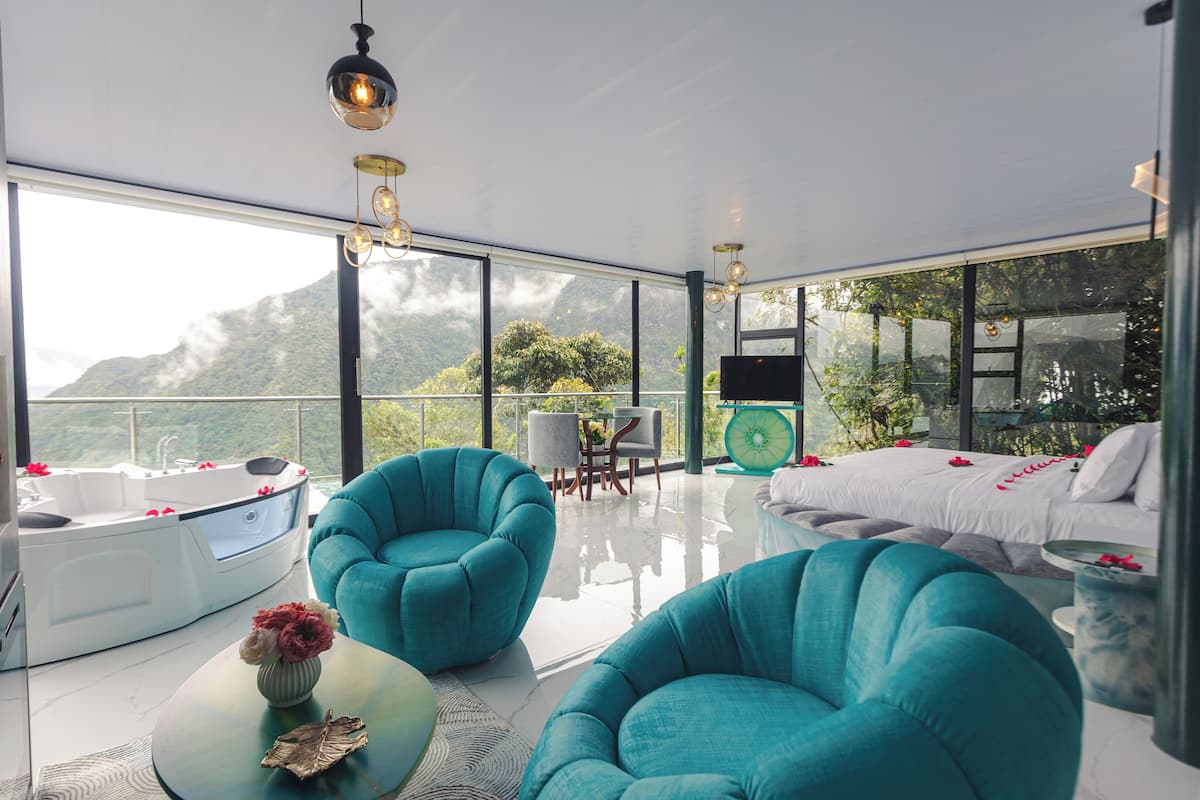 Ein modernes Schlafzimmer mit einem großen Bett, zwei türkisfarbenen Sesseln, einem weißen Sofa und einem verglasten Balkon mit Bergblick.