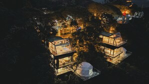Deux cabanes surélevées, dotées d’un toit en verre et de murs transparents, entourées d’arbres et éclairées par des lumières chaleureuses.
