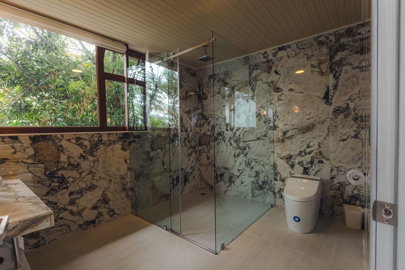 A bathroom with a glass shower enclosure, marble walls, and a toilet.