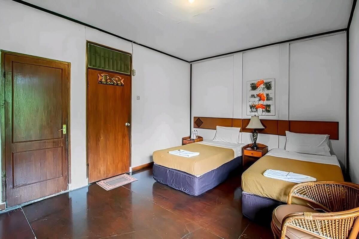 Une chambre d’hôtel avec deux lits, une porte en bois, un petit tapis, une œuvre d’art accrochée au mur et une chaise en osier.