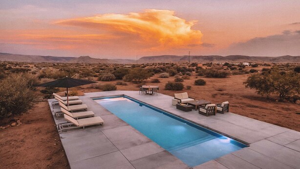 Piscina con sillones de descanso y una mesa, rodeada por un paisaje desértico y un cielo al atardecer.