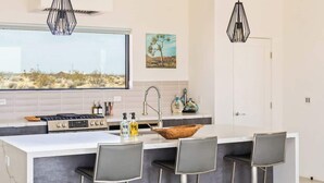 Una cocina moderna con una isla central, taburetes de bar, luces colgantes y un ventanal grande.