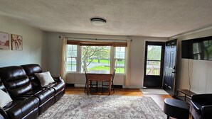 A living room with a black leather sofa, a dining table with chairs, a large window with curtains, a flat-screen TV mounted on the wall, and a door leading outside.