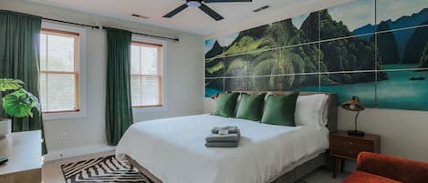 A bedroom with a bed, a ceiling fan, a nightstand, a lamp, a painting, and a window with curtains.
