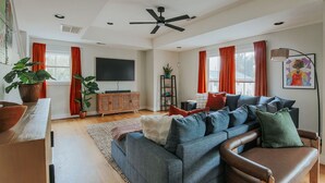 A spacious living room with a grey sofa, a wooden cabinet, a flat-screen TV, and a ceiling fan. - Oasis on 5th: walk to downtown Cville & UVA (Charlottesville,)