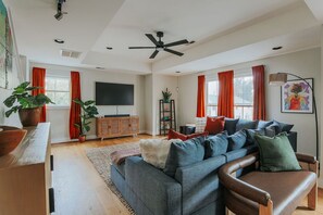 A spacious living room with a grey sofa, a wooden cabinet, a flat-screen TV, and a ceiling fan.