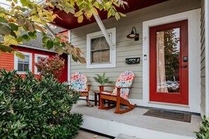 Une maison avec une porte rouge, une véranda avec deux fauteuils à bascule et une petite table, et une fenêtre au-dessus de la porte.