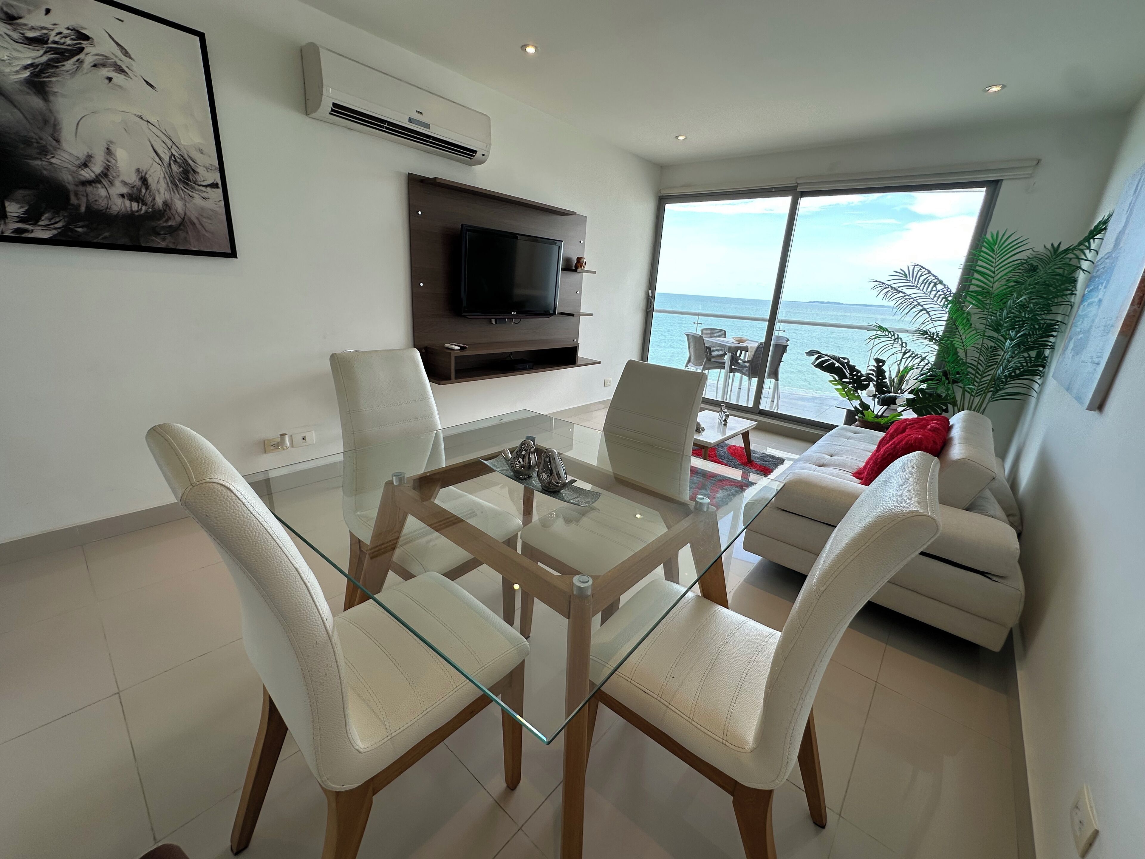 A modern living room with a glass dining table, white leather chairs, a sofa, and a TV mounted on the wall.