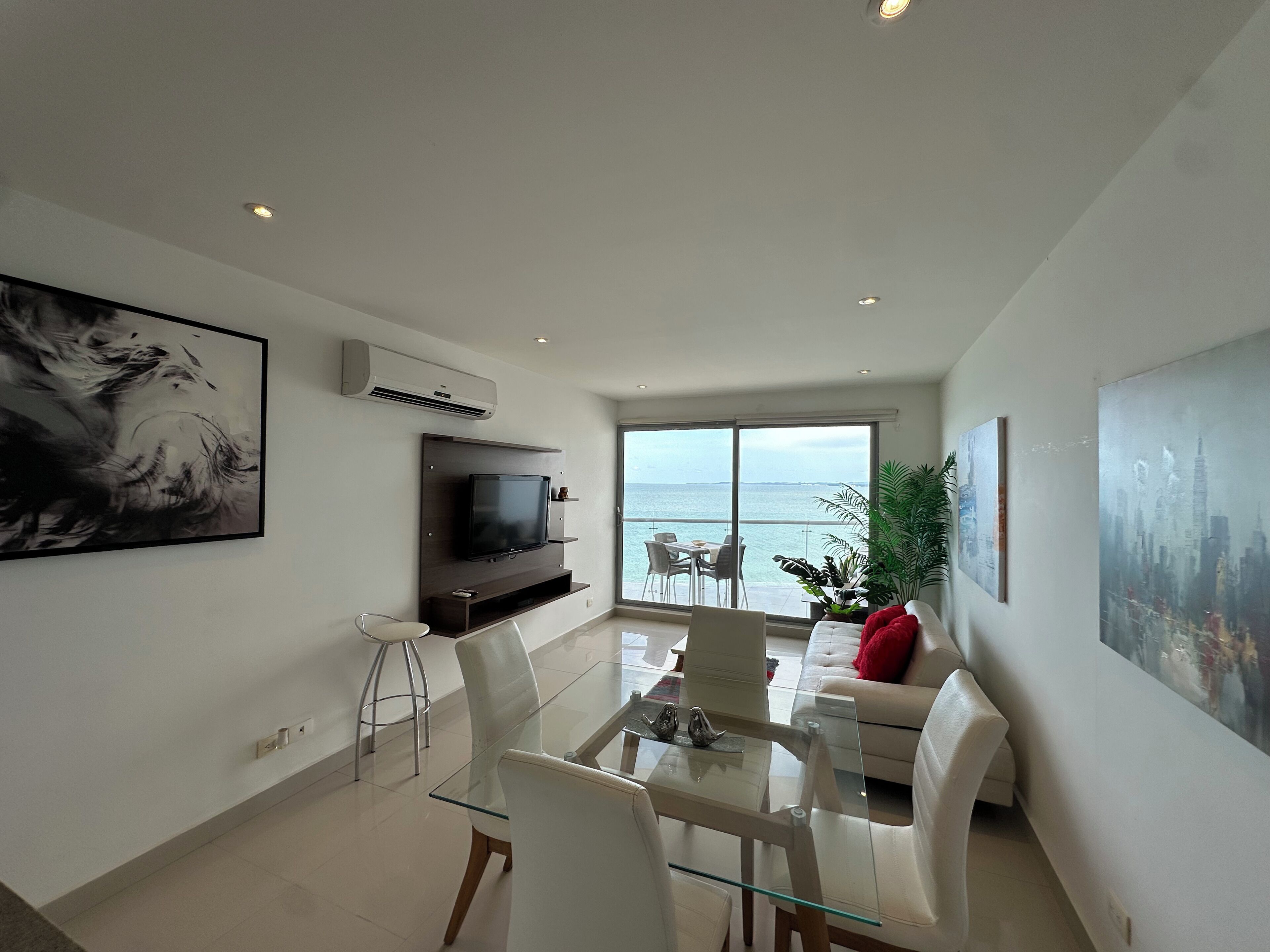 A modern living room with a glass dining table, white sofas, a flat-screen TV, and a large window overlooking the sea.