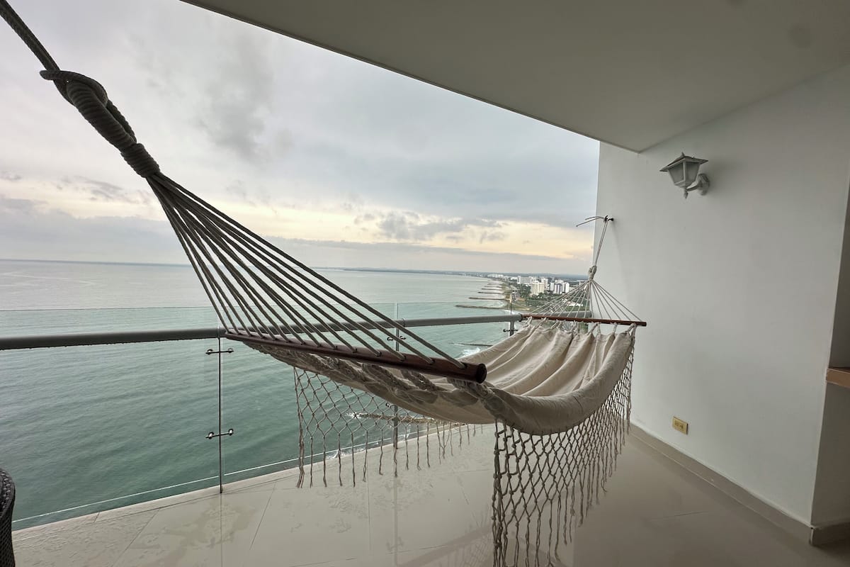 A hammock on a balcony with a view of the sea and buildings.