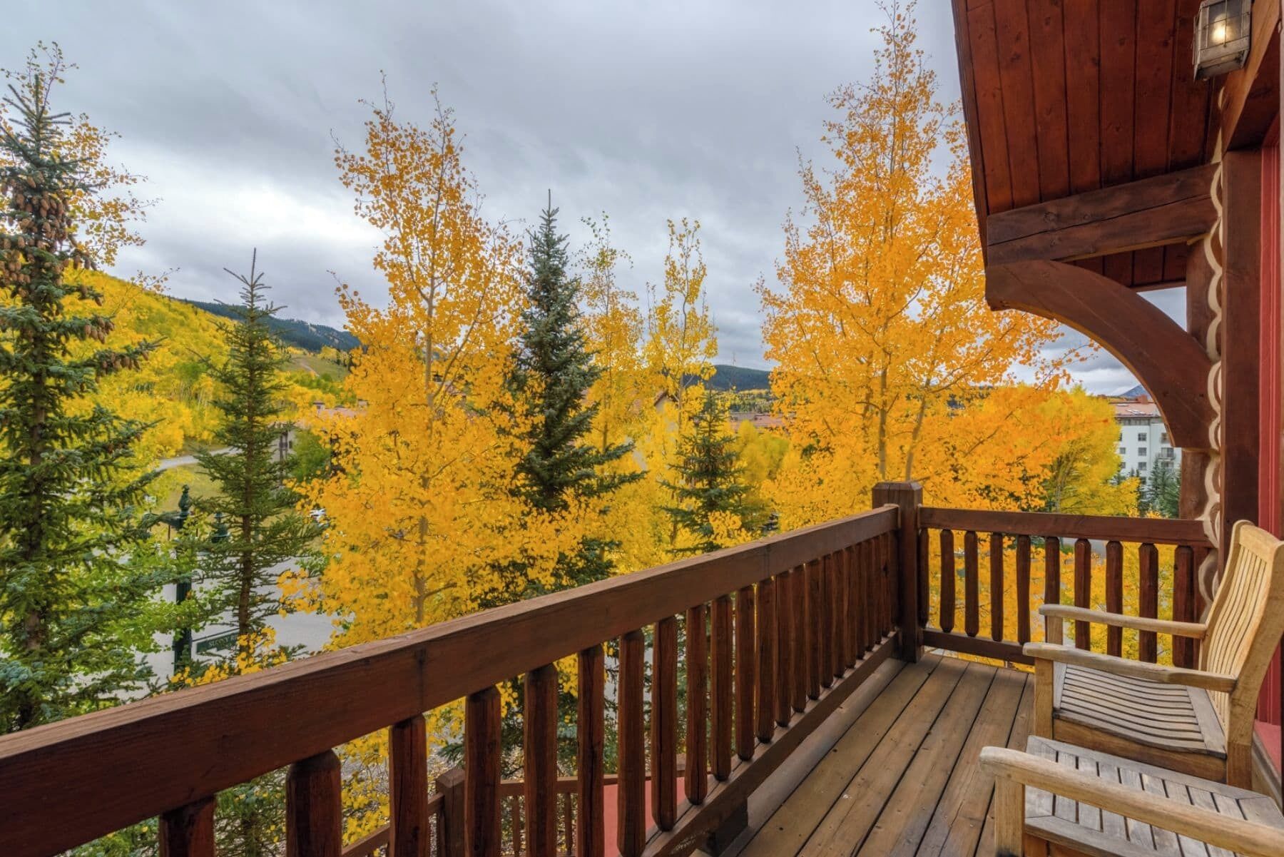 A wooden deck with a railing, overlooking a forest with yellow foliage and a view of buildings in the distance.