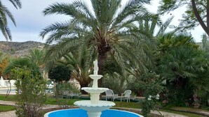 A stone fountain surrounded by a pool, with palm trees and a grassy area in the foreground. - Finca for events (family reunions, birthdays, weddings, seminars...) (Elda)