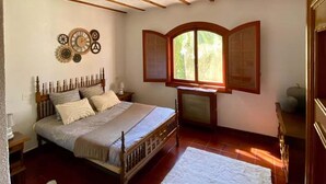 A bedroom with a wooden bed, a white rug, a window with a view of greenery, and a decorative wall piece. - Finca for events (family reunions, birthdays, weddings, seminars...) (Elda)