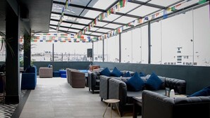 A rooftop lounge with leather sofas, a small round table, and a view of the cityscape through large windows. Flags are strung across the ceiling. - Hotel Ghangri (Kathmandu)