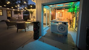An outdoor patio area with wicker furniture and a small table, a decorative black planter, and a tire on a platform. - The Sleepy Tiki - Game room, Tiki lounge, Fire pit, Fenced yard – Pet friendly! (Mason)