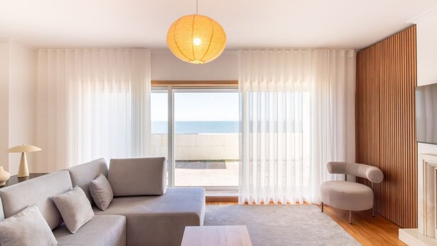 A modern living room with a sofa, a chair, a table, and a view of the sea through large windows.