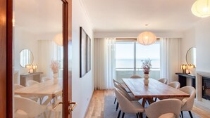 A dining area with a wooden table and chairs, a vase of flowers, and a view of the sea through large windows.