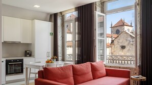 A modern living room with a red sofa, a dining table with chairs, and a kitchen area with white cabinets and appliances. A balcony with a wrought iron railing and a view of a historic building is visible through the window. - Bracara Home By Minhos Guest (Braga)