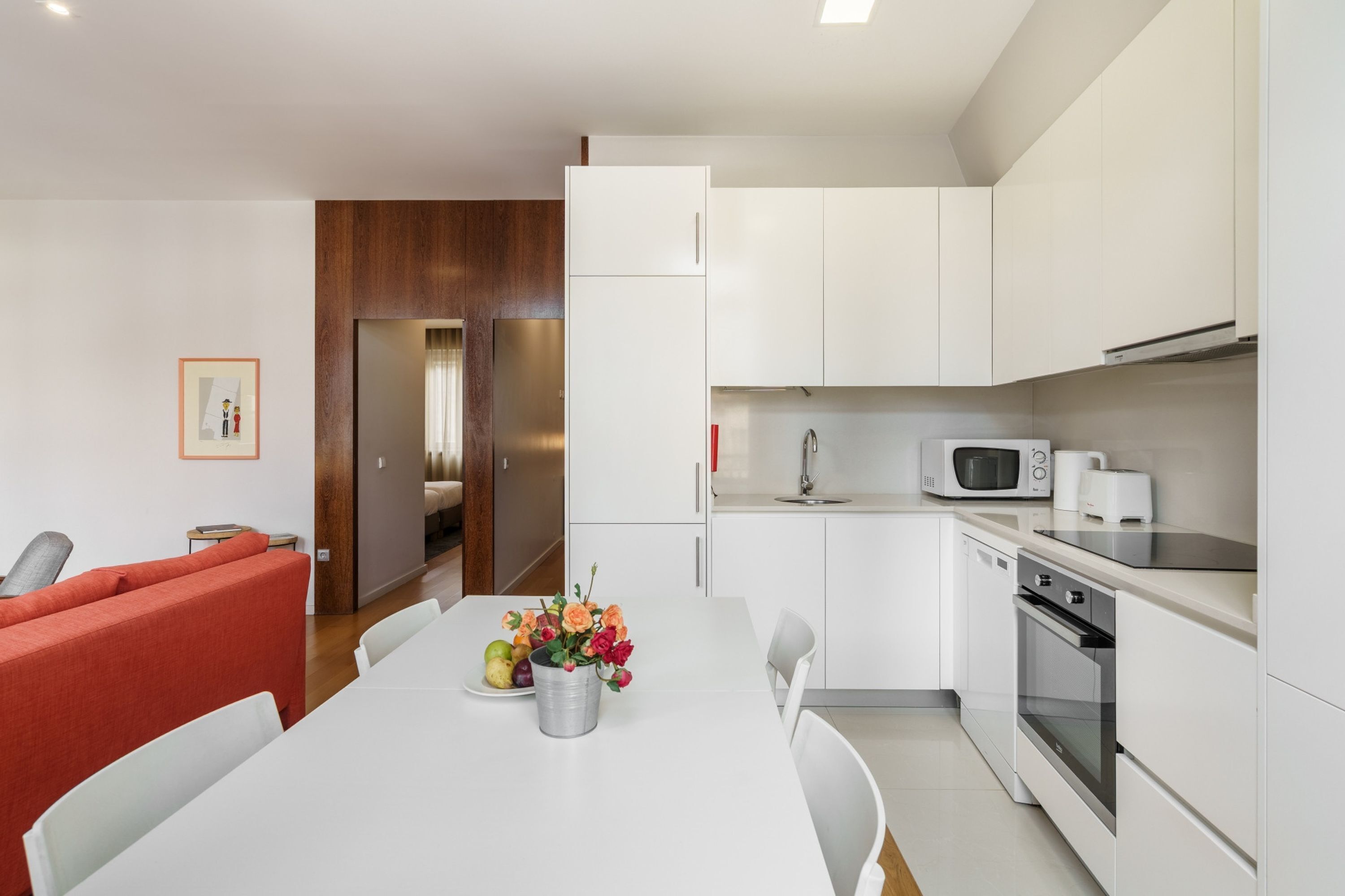 A modern kitchen with white cabinets, a microwave, and an oven. There is a dining table with chairs and a red sofa in the adjacent living area.