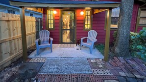 Un patio couvert avec deux fauteuils Adirondack, une clôture en bois et un bâtiment rouge doté d’un toit à pignon.