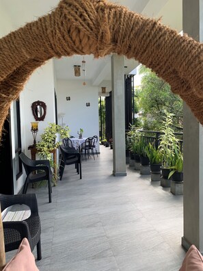 A covered outdoor area with a dining table and chairs, surrounded by potted plants and a view of greenery.