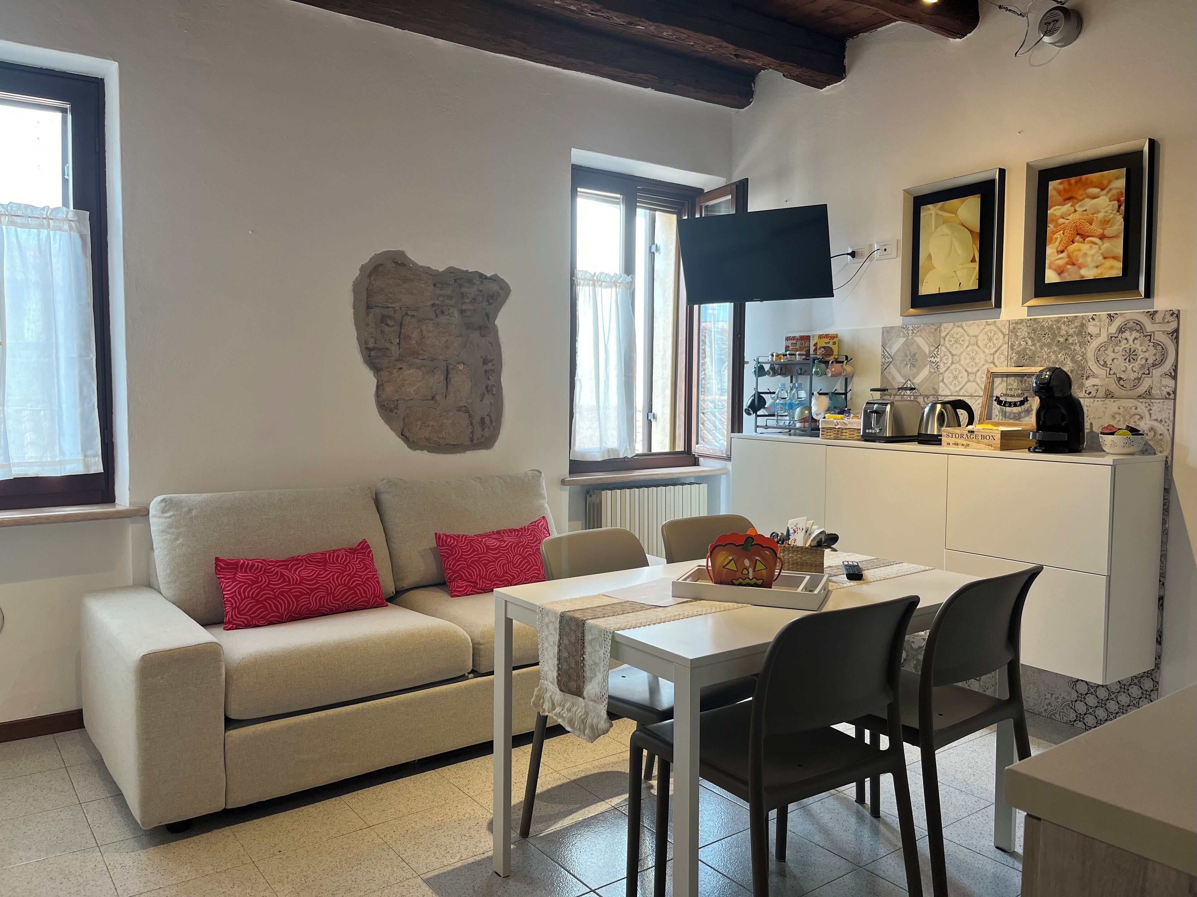 Ein gemütliches Wohnzimmer mit einer Couch, einem Esstisch und Stühlen. Im Hintergrund ist eine Küchenzeile mit einer Kaffeemaschine und einem Fernseher zu sehen.