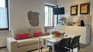Ein gemütliches Wohnzimmer mit einer Couch, einem Esstisch und Stühlen. Im Hintergrund ist eine Küchenzeile mit einer Kaffeemaschine und einem Fernseher zu sehen.