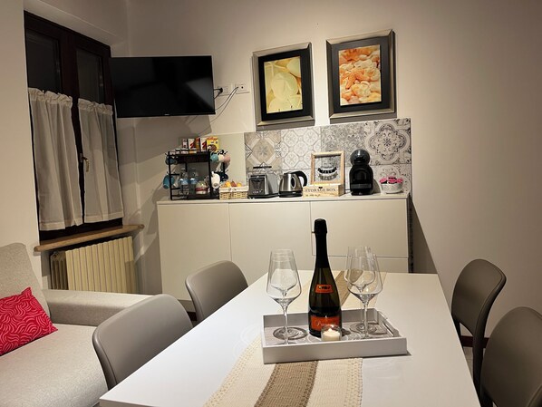 Un comedor con una mesa preparada para dos personas, una botella de vino y dos copas. Se ve una cocina con hervidor, tostadora y cafetera.