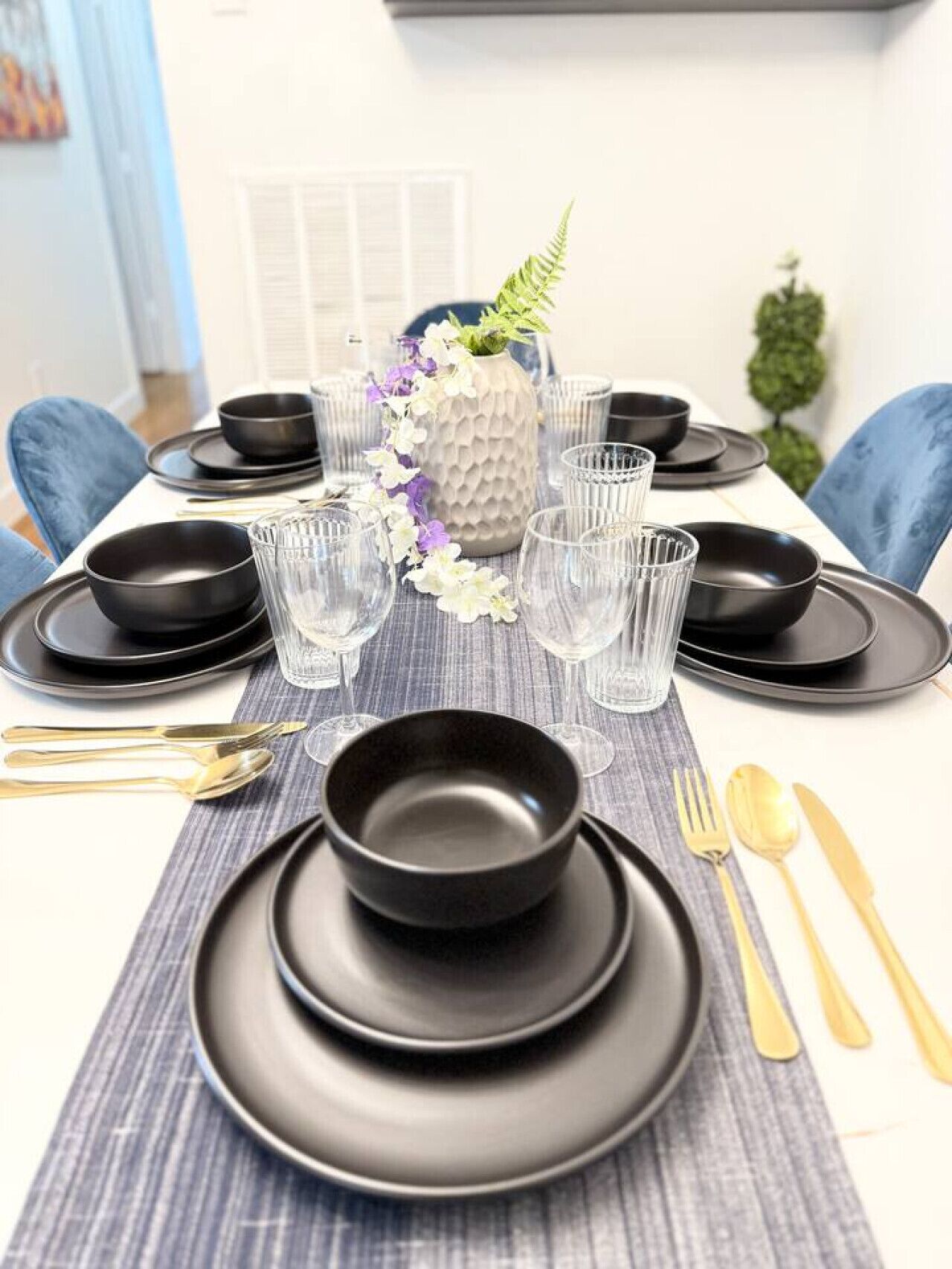 A dining table set with black plates, bowls, and clear glasses, centered with a decorative vase.