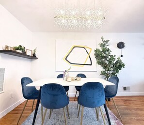 Een eetruimte met een witte tafel, blauwe stoelen, een kroonluchter, een geometrisch kunstwerk en een plant.