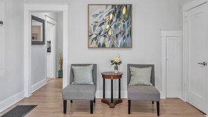 Ein Flur mit zwei grauen Sesseln, einem kleinen runden Tisch mit Blumenvase und einem großen abstrakten Gemälde an der Wand.