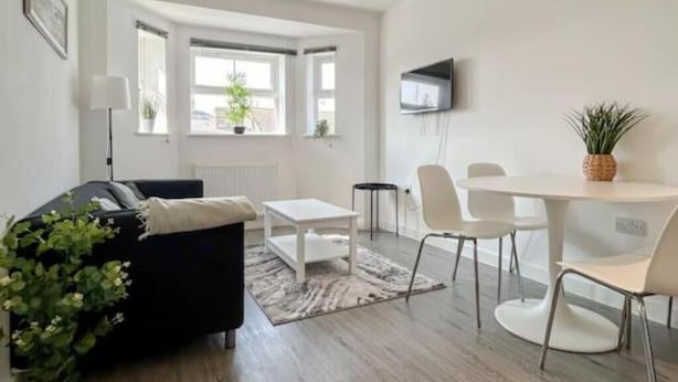 Un salon moderne avec un canapé noir, une table basse blanche et un coin repas comprenant des chaises et une table blanches. Un téléviseur est fixé au mur et une plante est posée sur la table du coin repas.